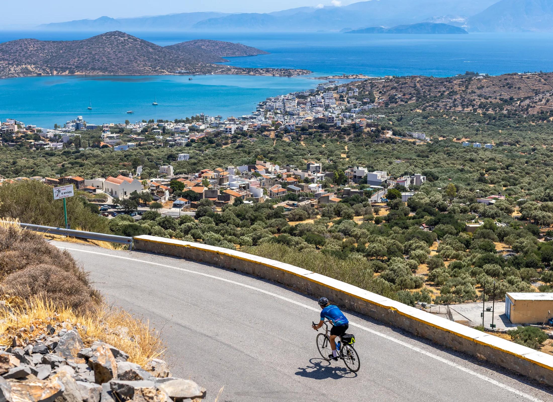 A cyclist riding down a winding road on a hillside overlooking the bay and coastal town of Elounda, Crete.