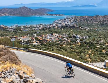 A cyclist riding down a winding road on a hillside overlooking the bay and coastal town of Elounda, Crete.