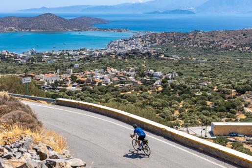 A cyclist riding down a winding road on a hillside overlooking the bay and coastal town of Elounda, Crete.