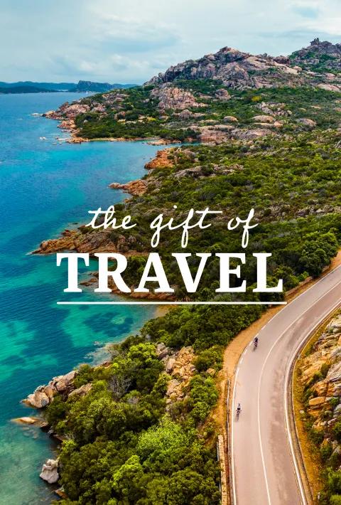 A coastal road winding along the turquoise waters and granite hills of Sardinia, Italy. Text overlay reads: The gift of travel. 