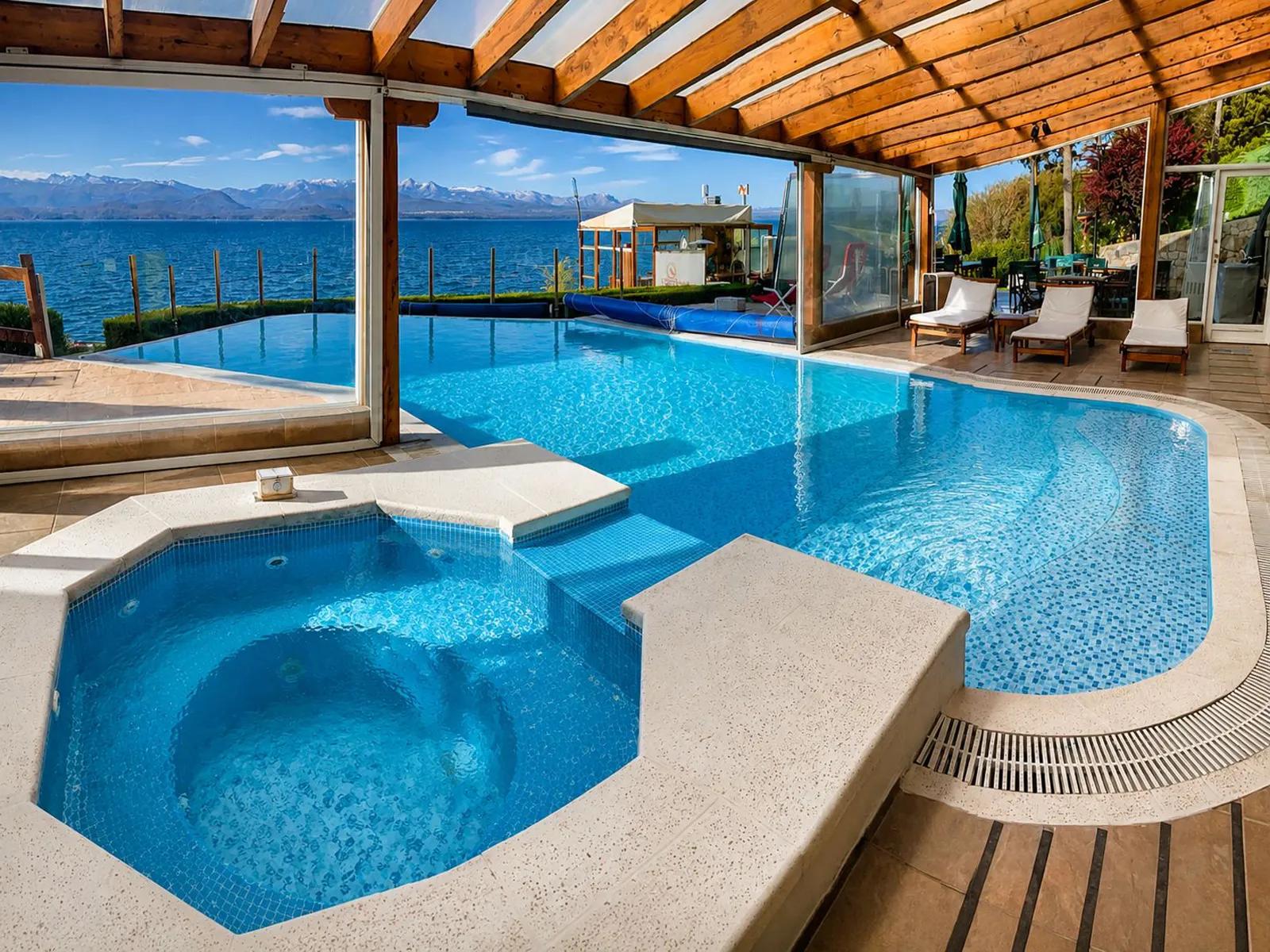An indoor-outdoor swimming pool and hot tub area with blue tiled water under a wooden-beamed roof at Sol del Nahuel Hotel & Spa in San Carlos de Bariloche, Argentina, featuring large glass windows offering scenic views of Lake Nahuel Huapi and distant snow-capped mountains.