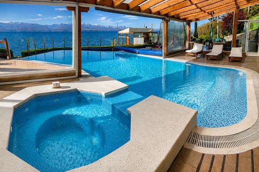 An indoor-outdoor swimming pool and hot tub area with blue tiled water under a wooden-beamed roof at Sol del Nahuel Hotel & Spa in San Carlos de Bariloche, Argentina, featuring large glass windows offering scenic views of Lake Nahuel Huapi and distant snow-capped mountains.