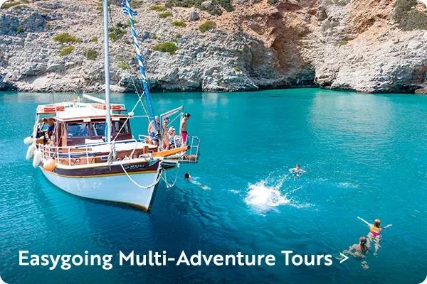 A wooden boat anchored in clear turquoise water near rocky cliffs, with people on board and several people swimming in the sea. In the lower left corner, bold white text reads "Easygoing Multi-Adventure Tours >".