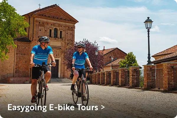 Two cyclists wearing matching blue cycling jerseys and helmets riding e-bikes side-by-side on a cobblestone road in a European town, with historic brick buildings and a church in the background. In the bottom left corner, white text reads "Easygoing E-bike Tours >".