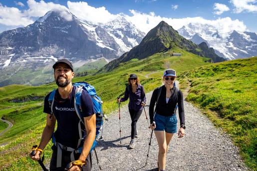 Three hikers on a scenic, gravel hiking trail in the Swiss Alps, surrounded by lush green hillsides. and dramatic, snow-dusted peaks.