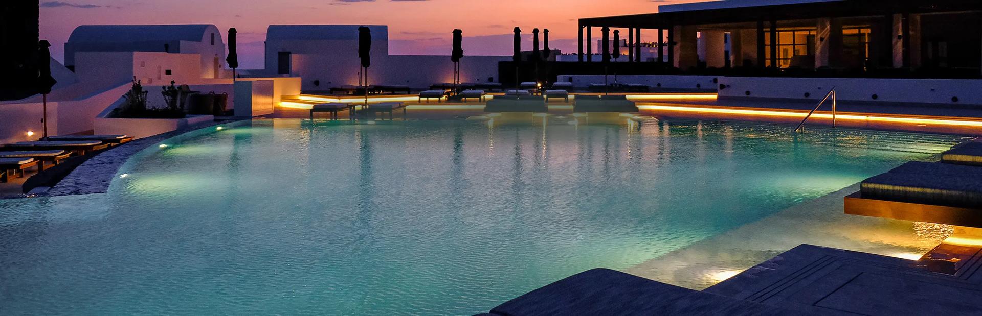 An illuminated swimming pool at twilight surrounded by lounge chairs, patio umbrellas, and white whitewashed buildings set against a warm sunset sky.