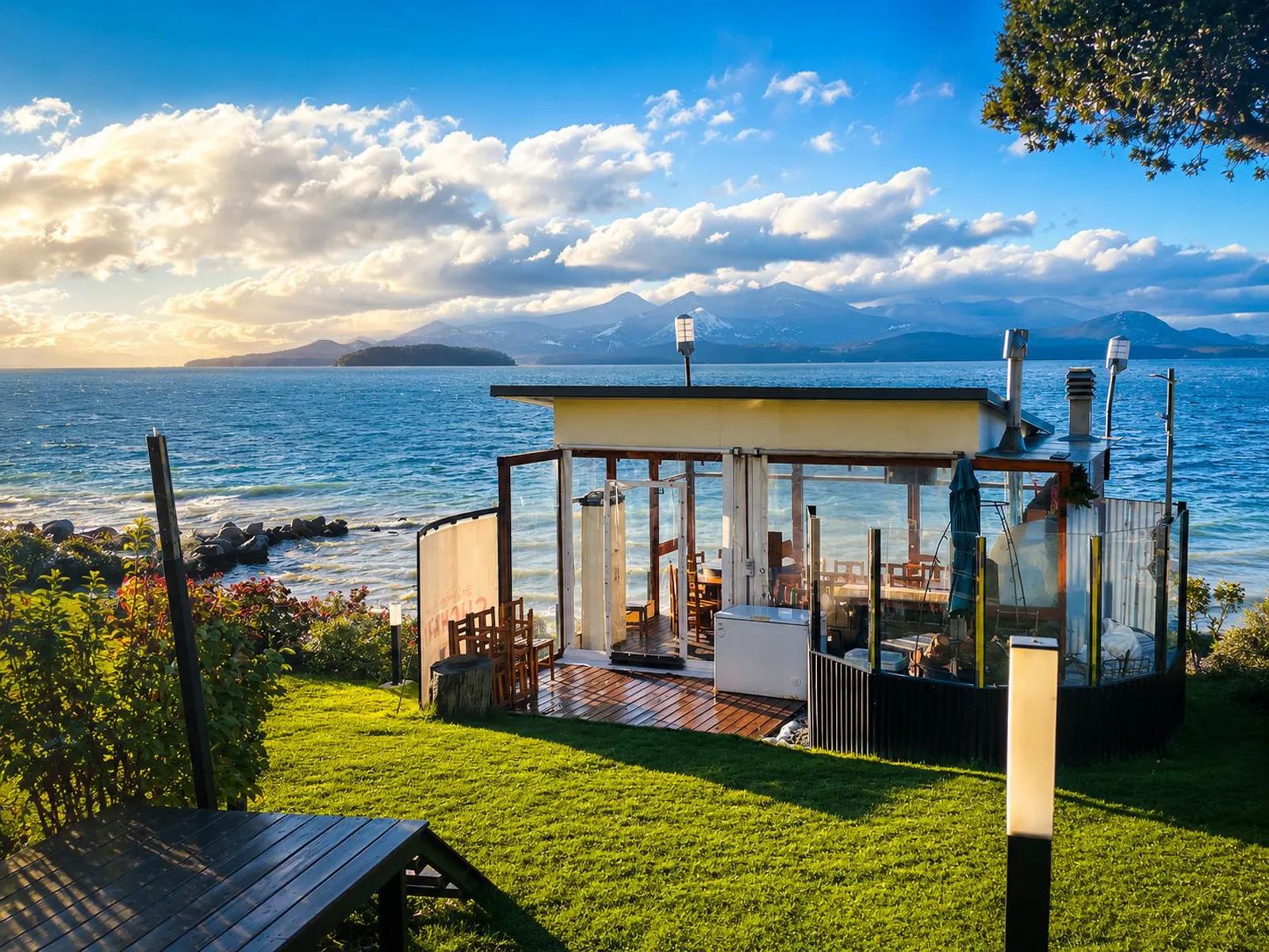 A glass-enclosed outdoor dining pavilion on a grassy slope at Sol del Nahuel Hotel & Spa in San Carlos de Bariloche, Argentina, offering scenic waterfront views of Lake Nahuel Huapi and surrounding mountains under a partly cloudy sky.
