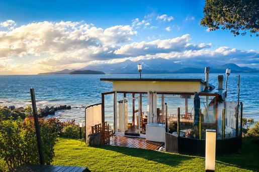 A glass-enclosed outdoor dining pavilion on a grassy slope at Sol del Nahuel Hotel & Spa in San Carlos de Bariloche, Argentina, offering scenic waterfront views of Lake Nahuel Huapi and surrounding mountains under a partly cloudy sky.