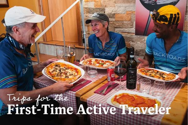 Three Backroads cycling guests sitting at a cozy restaurant table, each holding a personal-sized pizza on an oval plate next to a bottle of red wine. Text overlay reads: Trips for the First-Time Active Traveler. 