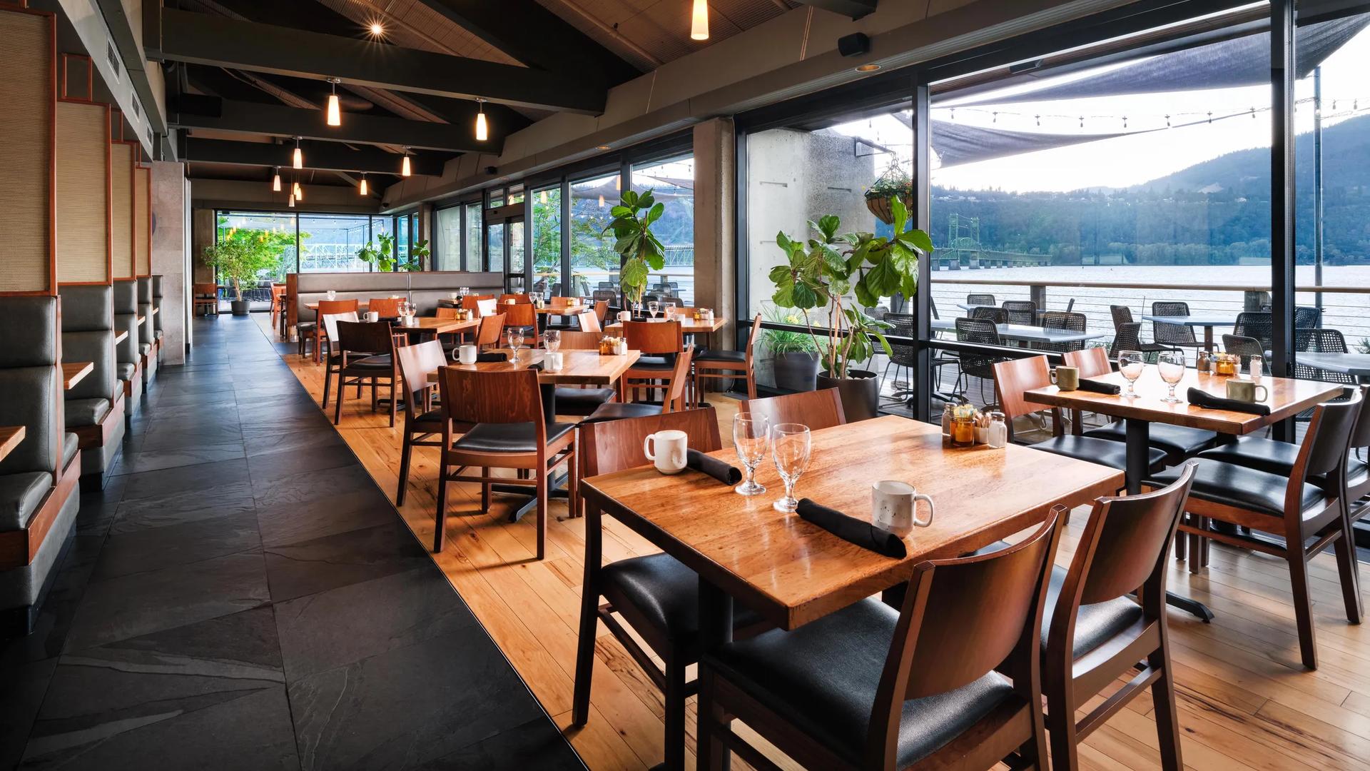 A spacious indoor restaurant dining room at the Hood River Inn with wooden tables, warm lighting, and floor-to-ceiling windows overlooking a river and mountains.