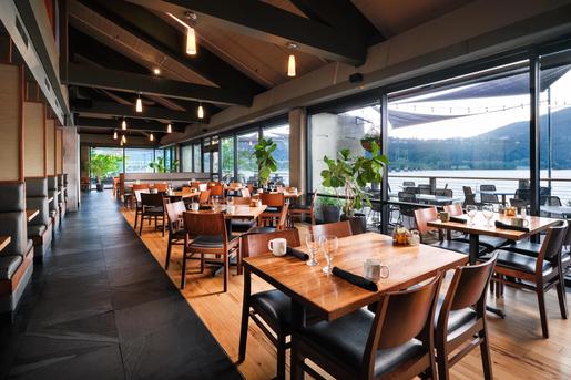 A spacious indoor restaurant dining room at the Hood River Inn with wooden tables, warm lighting, and floor-to-ceiling windows overlooking a river and mountains.