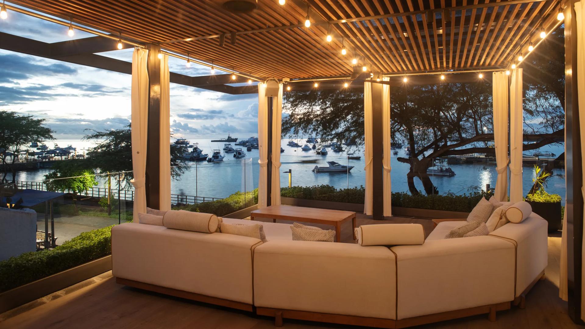 An outdoor seating lounge area at Hotel Indigo Galapagos, features a plush white couch and a modern wooden slat pergola overlooking a scenic harbor.