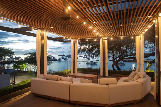 An outdoor seating lounge area at Hotel Indigo Galapagos, features a plush white couch and a modern wooden slat pergola overlooking a scenic harbor.
