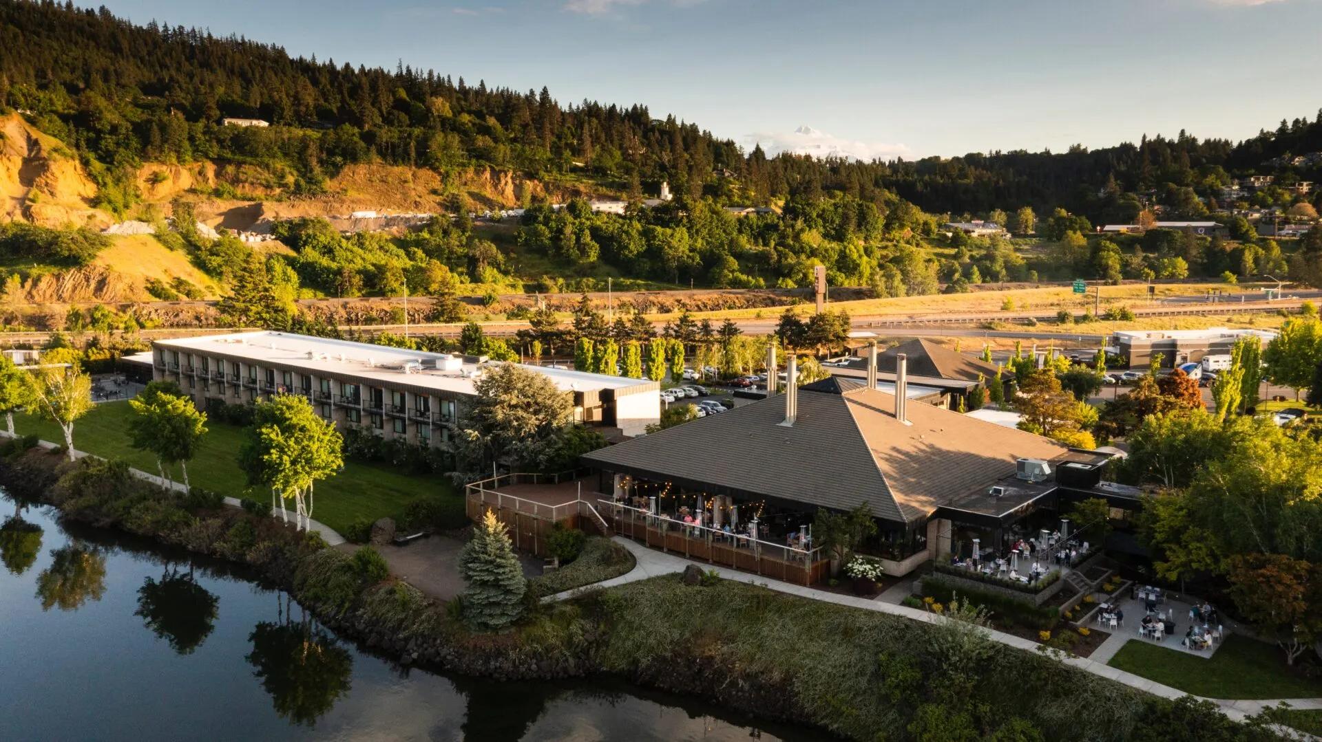 An aerial view of a riverside resort complex with outdoor patio seating, a grassy lawn, and forested hills in the background under warm sunlight.