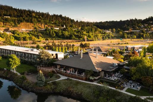 An aerial view of a riverside resort complex with outdoor patio seating, a grassy lawn, and forested hills in the background under warm sunlight.