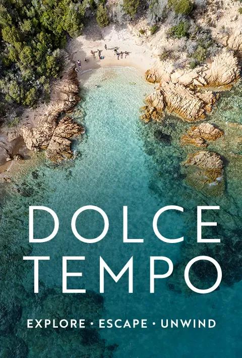 An aerial overhead view of a secluded beach inlet with clear turquoise water, sandy shore with a few people, and rocky cliffs surrounded by greenery. Overlaid on the lower half of the image in large white text is "DOLCE TEMPO" above the subtitle "EXPLORE • ESCAPE • UNWIND"