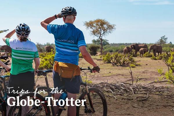 Two cyclists wearing branded Backroads apparel looking out at a group of wild elephants in an African savanna landscape. Text overlay reads: Trips for the Globe-Trotter.