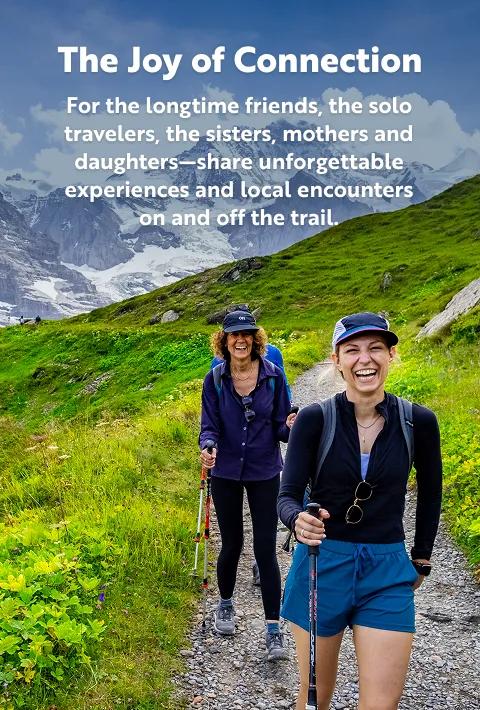 Panoramic landscape view of two smiling women hiking along a gravel trail in vibrant, lush green mountains under a blue sky. White text overlay on the left reads, "The Joy of Connection" with the subtext, "For the longtime friends, the solo travelers, the sisters, mothers and daughters—share unforgettable experiences and local encounters on and off the trail."