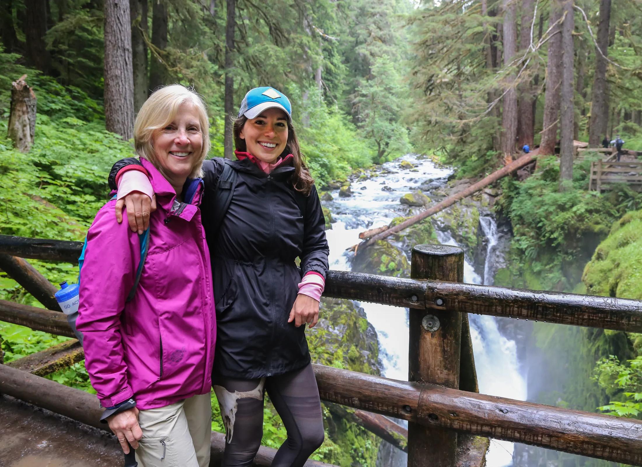 Two women smiling together on a wooden viewing platform along a scenic hiking trail. The woman on the left has short blonde hair and is wearing a pink jacket, while the woman on the right has long dark hair, a blue cap, and a dark jacket. They are standing in front of a rushing river and waterfall surrounded by a lush green forest.