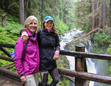 Two women smiling together on a wooden viewing platform along a scenic hiking trail. The woman on the left has short blonde hair and is wearing a pink jacket, while the woman on the right has long dark hair, a blue cap, and a dark jacket. They are standing in front of a rushing river and waterfall surrounded by a lush green forest.