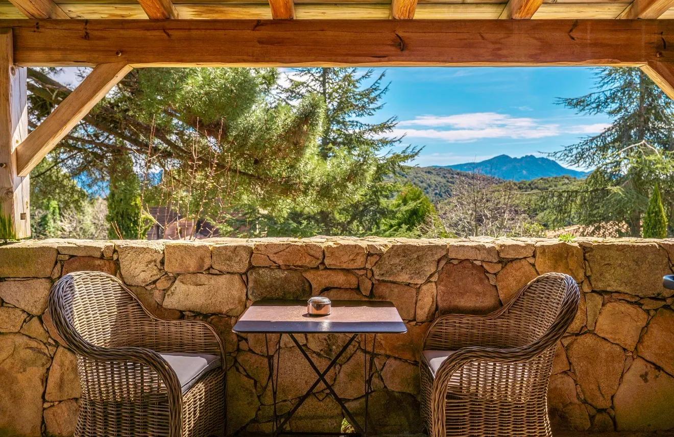 A terrace with traditional dry-stone walls, rustic wooden framing, and cozy wicker armchairs with a view overlooking dense pine forests and mountain peaks at A Pignata, in Levie, Corsica, France.