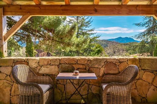 A terrace with traditional dry-stone walls, rustic wooden framing, and cozy wicker armchairs with a view overlooking dense pine forests and mountain peaks at A Pignata, in Levie, Corsica, France.