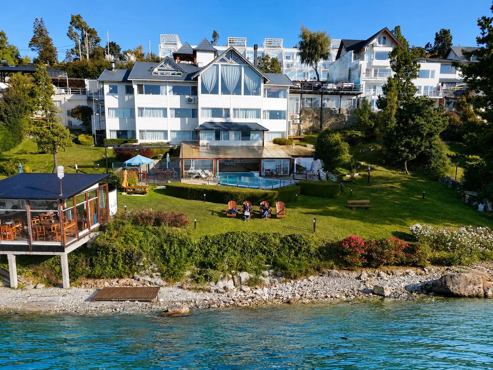 An elevated view of the waterfront exterior of Sol del Nahuel Hotel & Spa in San Carlos de Bariloche, Argentina, featuring a multi-story white alpine-style building set on a grassy hill leading down to a rocky shore on Lake Nahuel Huapi, complete with an outdoor swimming pool, patio seating, and lush green trees.