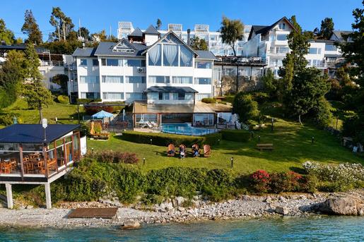 An elevated view of the waterfront exterior of Sol del Nahuel Hotel & Spa in San Carlos de Bariloche, Argentina, featuring a multi-story white alpine-style building set on a grassy hill leading down to a rocky shore on Lake Nahuel Huapi, complete with an outdoor swimming pool, patio seating, and lush green trees.