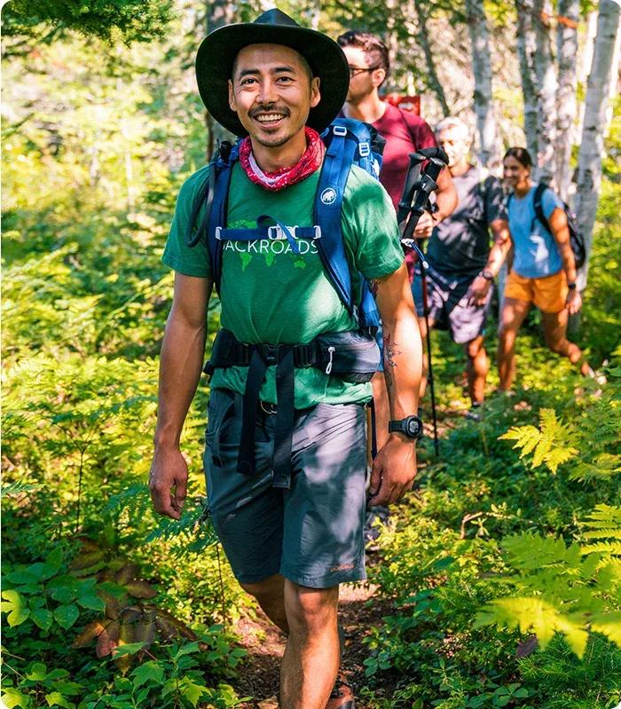 A smiling male hiking guide wearing a wide-brimmed hat, green shirt, backpack, and red neck gaiter leading a group of hikers along a lush, sunlit forest trail.