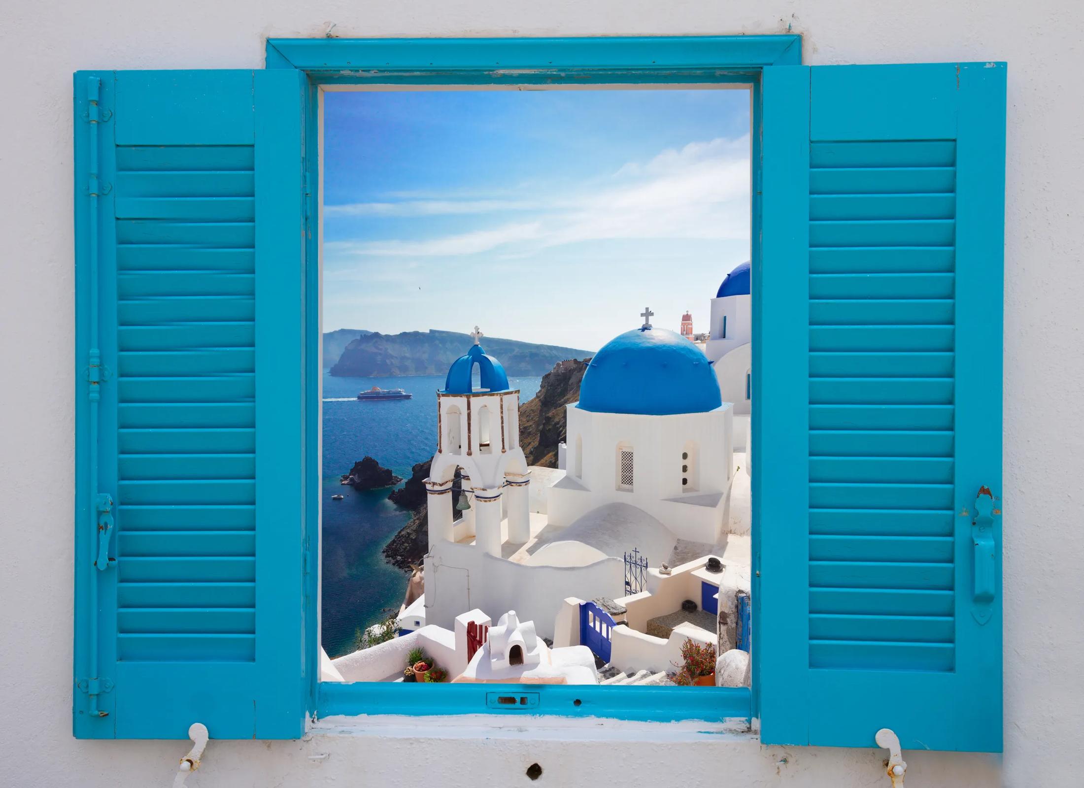 A view through open bright blue window shutters framing whitewashed, blue-domed churches on a cliffside overlooking the Aegean Sea in Santorini, Greece.