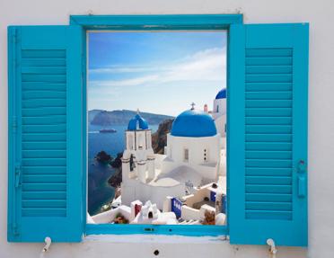 A view through open bright blue window shutters framing whitewashed, blue-domed churches on a cliffside overlooking the Aegean Sea in Santorini, Greece.