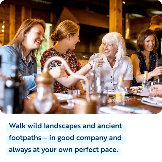 Four women sit around a dining table inside a warm, rustic room with wooden accents, laughing and talking together over drinks. Below the photo, dark blue text on a white background reads, "Walk wild landscapes and ancient footpaths &ndash; in good company and always at your own perfect pace."