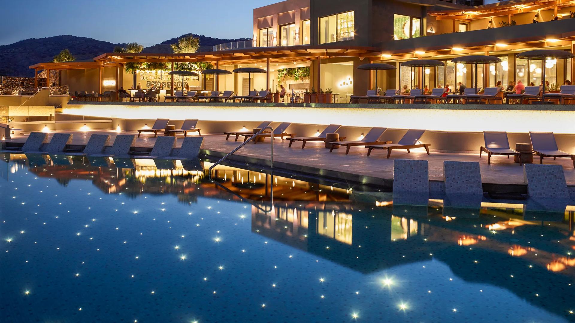 The outdoor swimming pool at twilight with tiny lights at the bottom resembling stars, with lounge chairs and mountains in the background at the Cayo Exclusive Resort & Spa in Plaka, Greece.