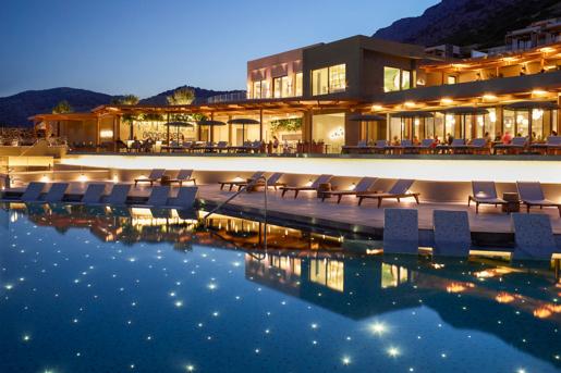 The outdoor swimming pool at twilight with tiny lights at the bottom resembling stars, with lounge chairs and mountains in the background at the Cayo Exclusive Resort & Spa in Plaka, Greece.