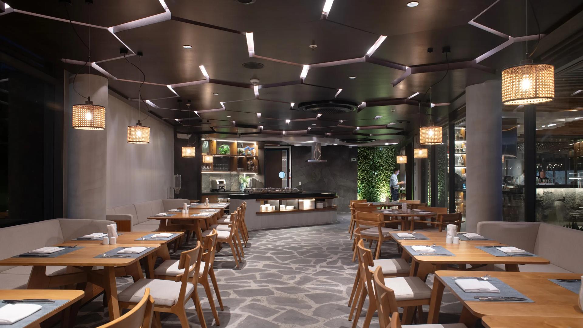 Darwin's Secret restaurant features a modern interior with light wood dining tables, cushioned chairs, grey flagstone flooring, and woven rattan pendant lights, located within the Hotel Indigo Galapagos on San Cristóbal Island.