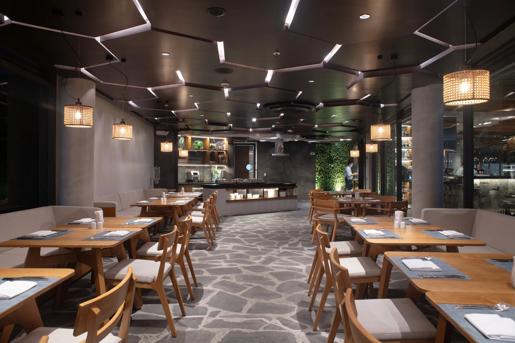 Darwin's Secret restaurant features a modern interior with light wood dining tables, cushioned chairs, grey flagstone flooring, and woven rattan pendant lights, located within the Hotel Indigo Galapagos on San Cristóbal Island.