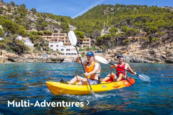 Two men in life vests paddling a yellow and orange tandem kayak on clear blue ocean water, with a rocky hillside and coastal buildings in the background and text reading "Multi-Adventures" overlaid in the bottom left corner.