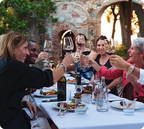 A group of smiling friends sitting around a white tablecloth dinner table outdoors near brick stone arches, raising their glasses for a toast over a meal and drinks.