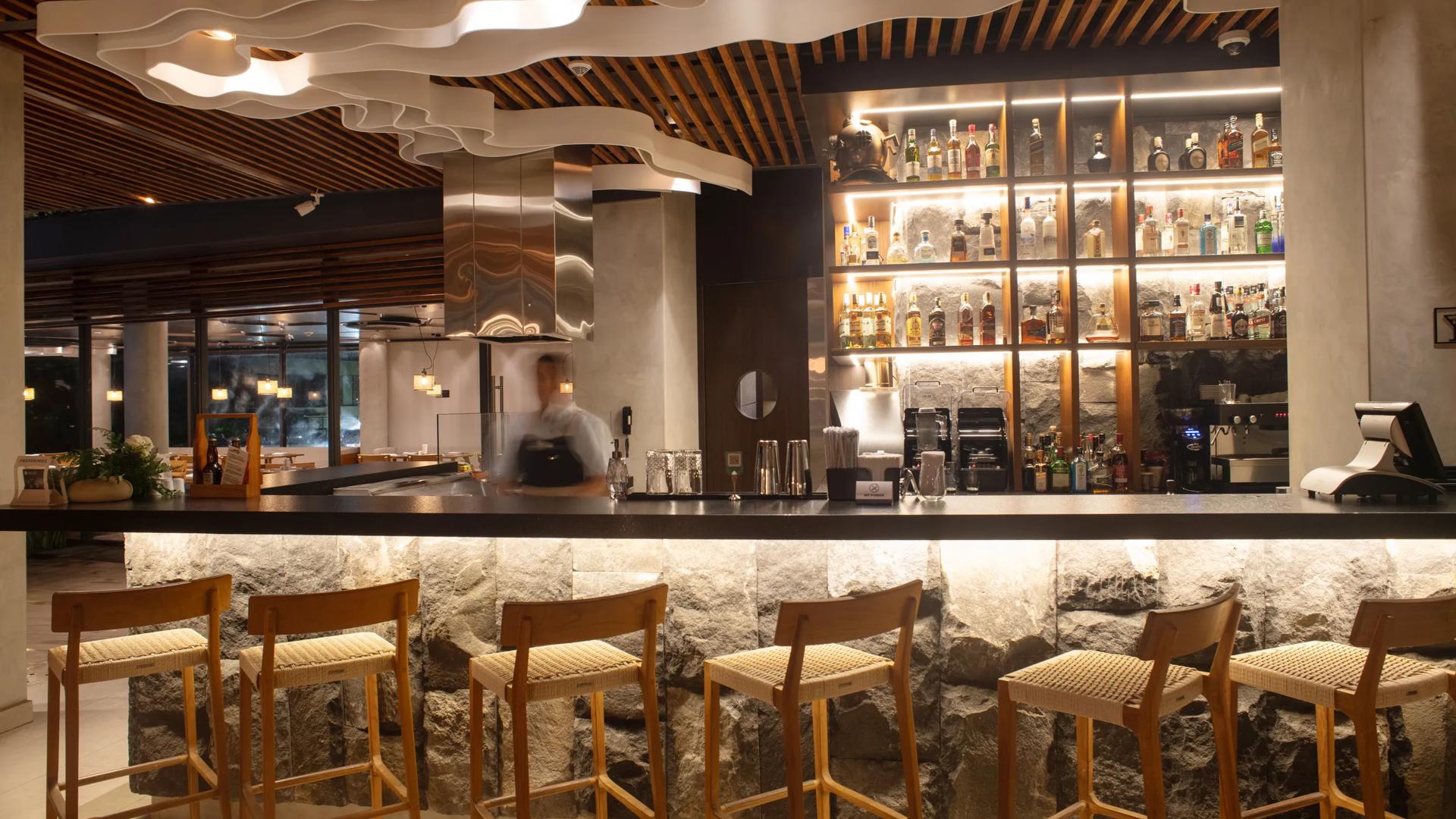 The modern bar inside Hotel Indigo Galapagos which features a wooden shelf with liquor, a wavy ceiling installation and wooden, tall bar chairs. 