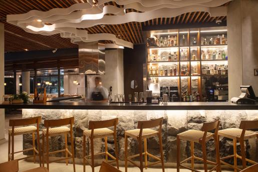 The modern bar inside Hotel Indigo Galapagos which features a wooden shelf with liquor, a wavy ceiling installation and wooden, tall bar chairs.