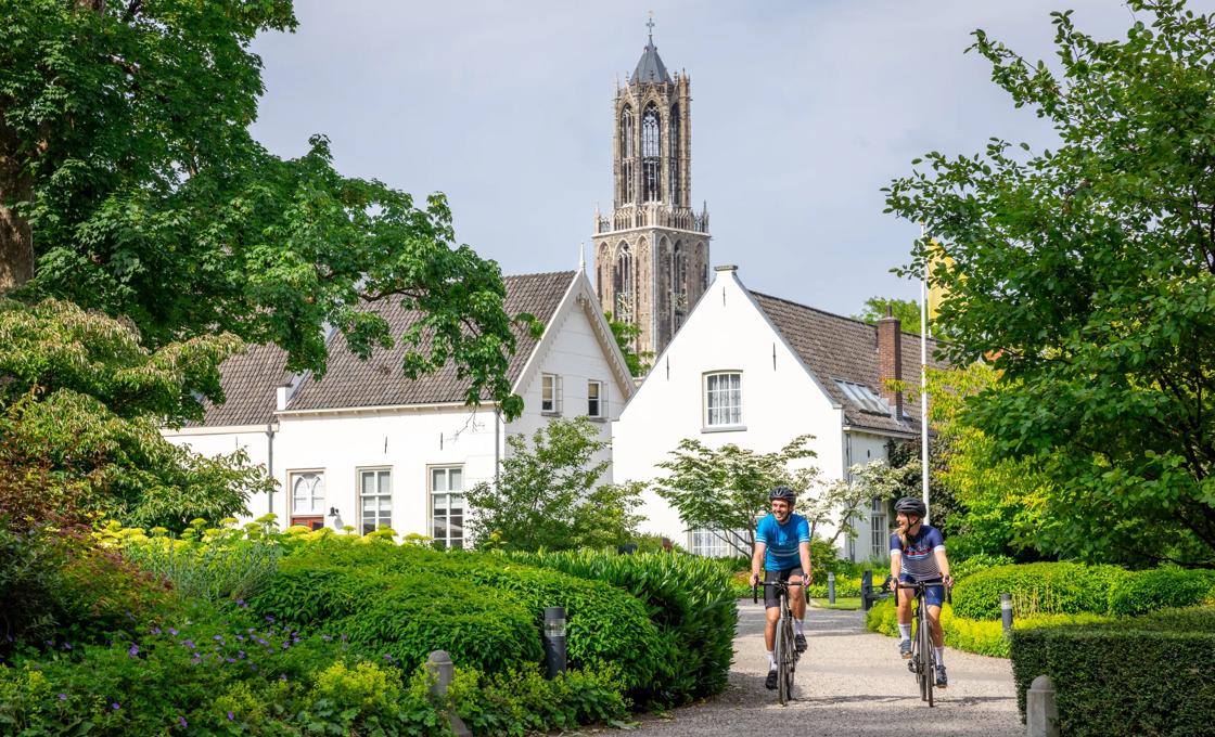Two cyclists ride in the lush landscape in front of a church in the Netherlands.