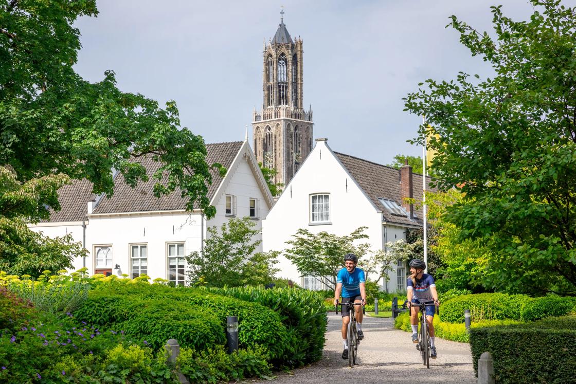 Two cyclists ride in the lush landscape in front of a church in the Netherlands.
