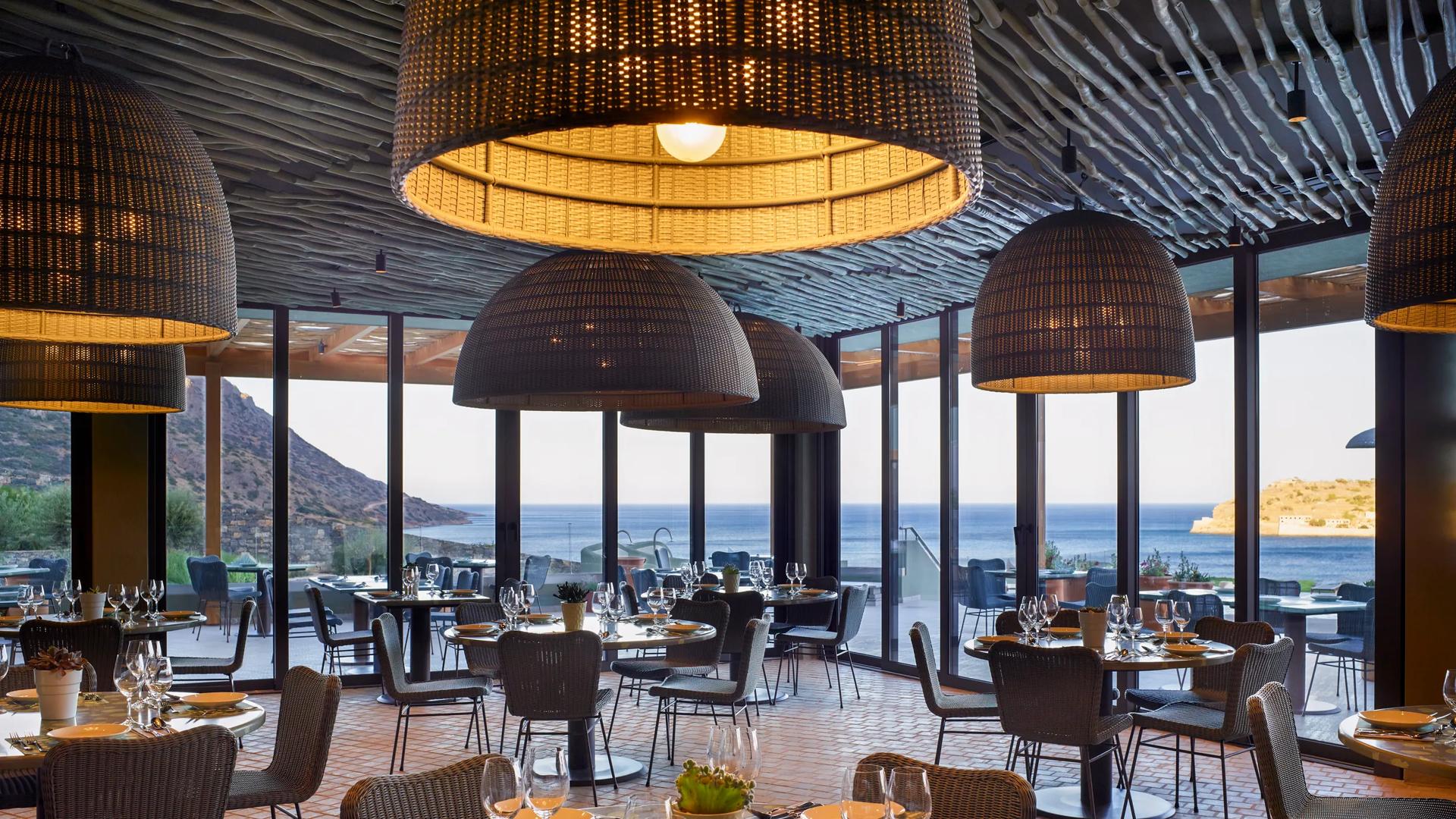 The Ambrosia Main Restaurant at the Cayo Exclusive Resort & Spa in Crete, featuring large woven pendant lights, woven chairs, and expansive floor-to-ceiling glass windows that open up to an outdoor patio.
