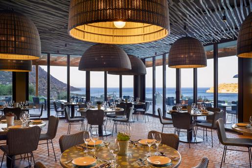 The Ambrosia Main Restaurant at the Cayo Exclusive Resort & Spa in Crete, featuring large woven pendant lights, woven chairs, and expansive floor-to-ceiling glass windows that open up to an outdoor patio.