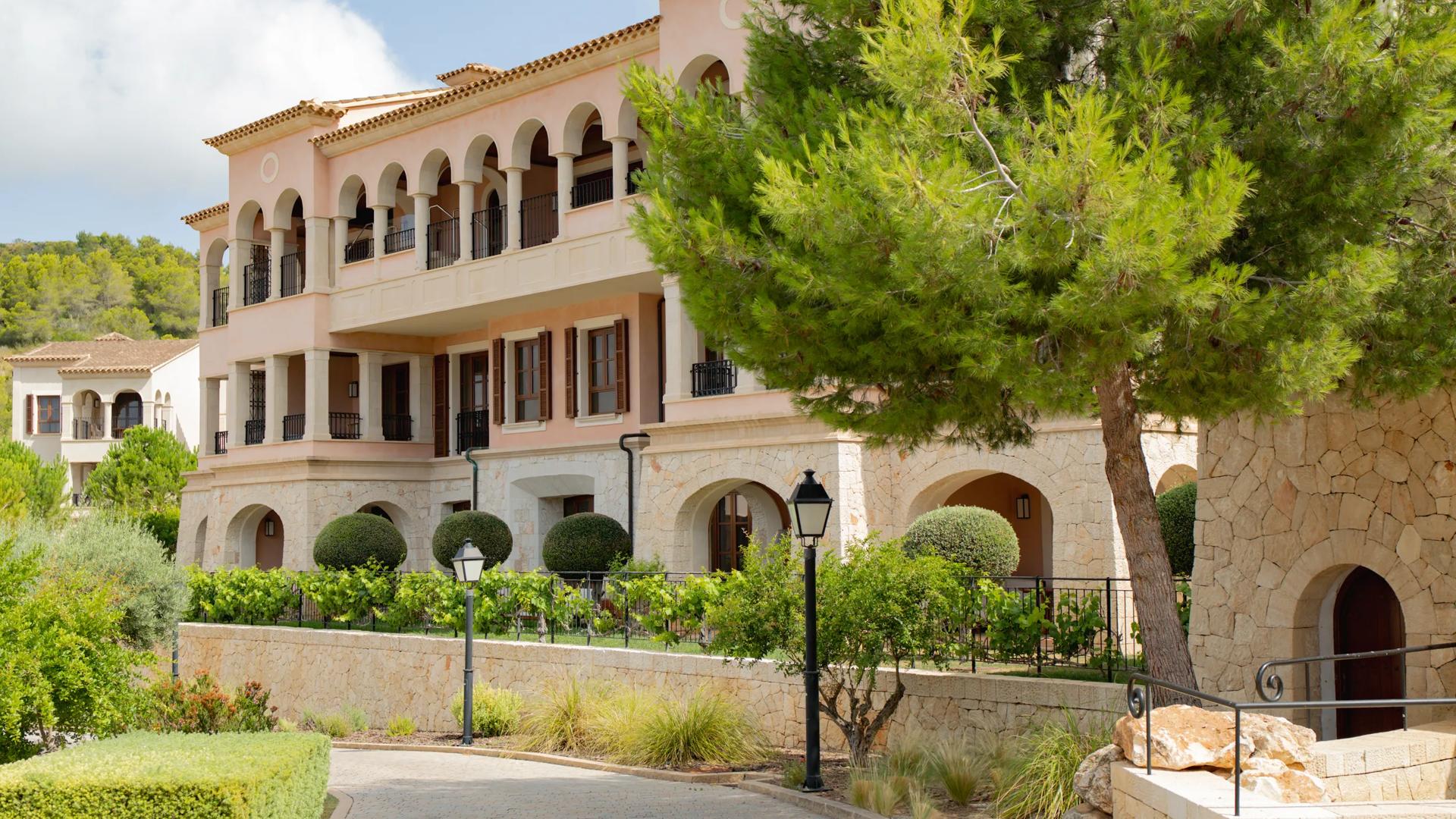 Exterior view of the Cap Vermell Grand Hotel, a Mediterranean-style hotel building with light pink walls, arched balconies, stone walls, and a large pine tree framing the foreground.
