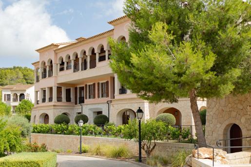 Exterior view of the Cap Vermell Grand Hotel, a Mediterranean-style hotel building with light pink walls, arched balconies, stone walls, and a large pine tree framing the foreground.