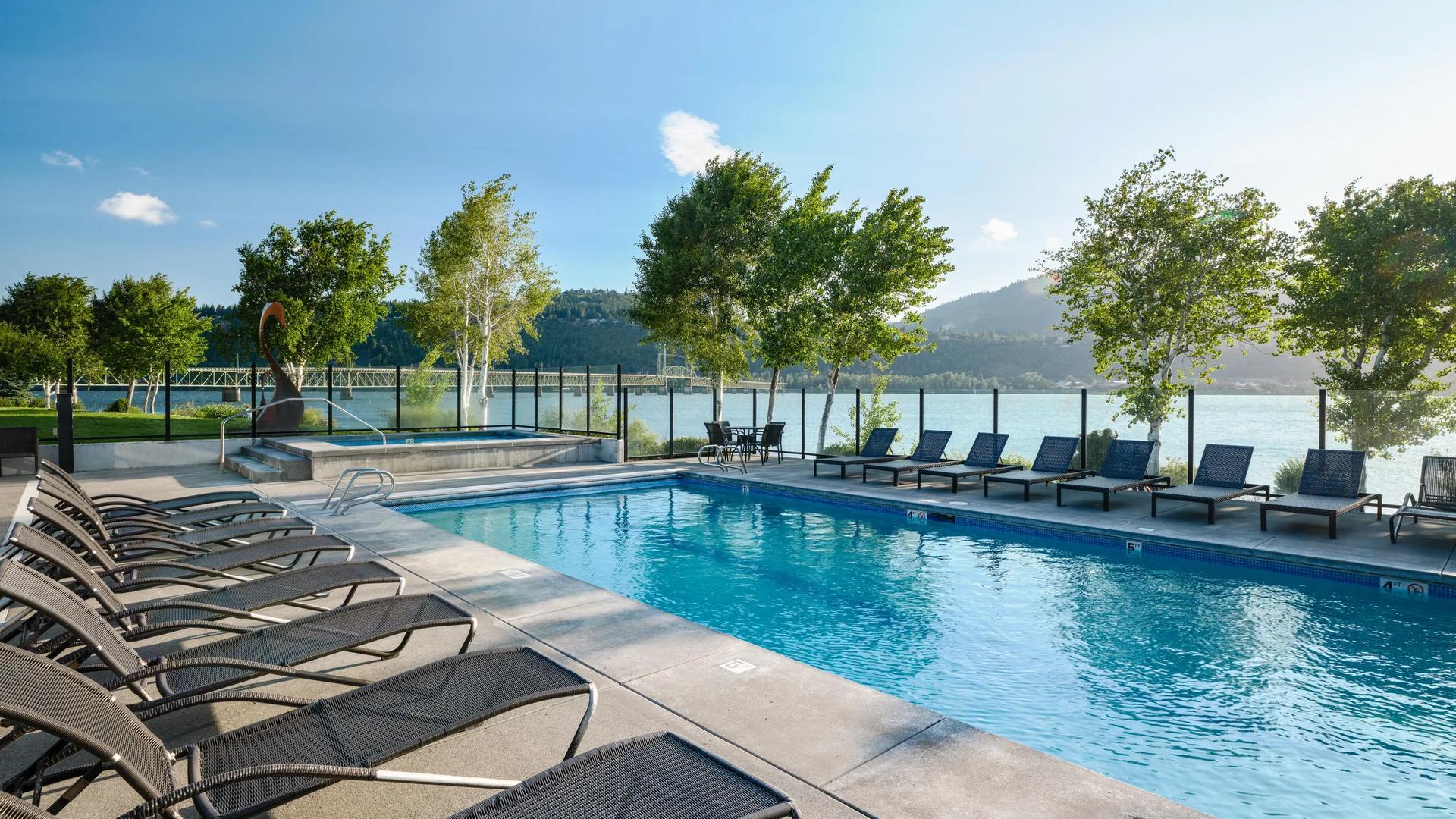 An outdoor swimming pool and hot tub area lined with lounge chairs at the Hood River Inn, overlooking a river and bridge on a sunny day.