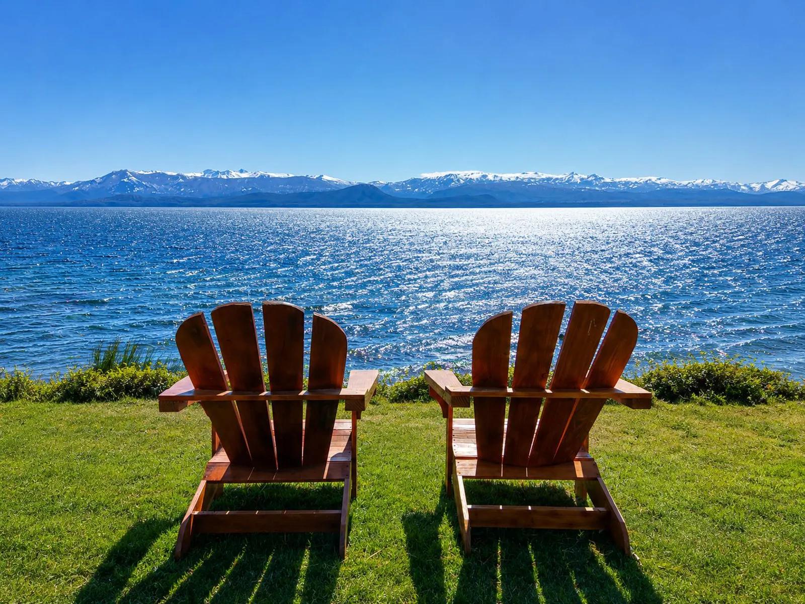 Two wooden Adirondack chairs sit on a grassy lawn at Sol del Nahuel Hotel & Spa in San Carlos de Bariloche, Argentina, facing a wide, bright blue lake under a clear sky, with snow-capped mountains stretching along the horizon in the background.