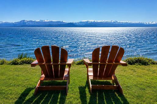 Two wooden Adirondack chairs sit on a grassy lawn at Sol del Nahuel Hotel & Spa in San Carlos de Bariloche, Argentina, facing a wide, bright blue lake under a clear sky, with snow-capped mountains stretching along the horizon in the background.