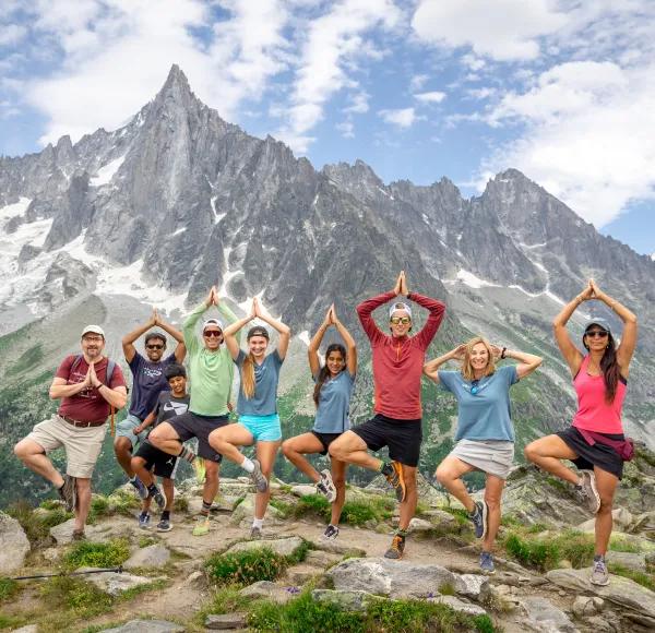 A diverse group of eight hikers posing in tree yoga poses on a rocky mountain trail with a dramatic, pointed rocky peak and mountain range in the background under a partly cloudy sky.